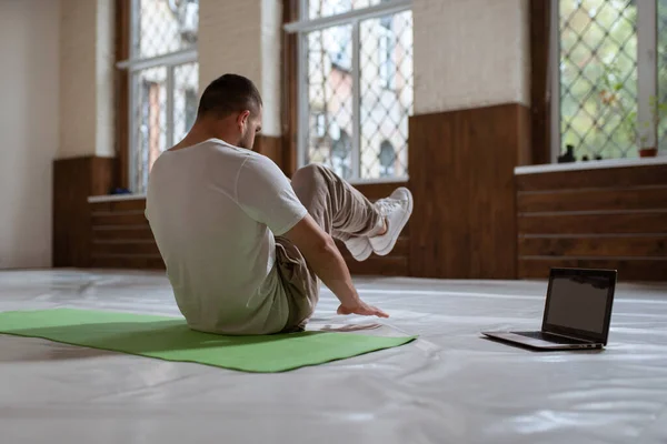 Vista posterior de un hombre guapo haciendo crujidos mientras ve videos de ejercicios en línea sobre el uso de la computadora portátil. Auto motivado joven calentamiento haciendo ejercicios especiales para los músculos abdominales — Foto de Stock