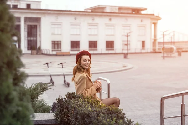 Belle femme de style français dans un manteau d'automne et beige se trouve sur un lit de fleurs marbrées avec partager ou louer des scooters électriques sur fond attendant sa date tenant tasse à café. Photo tonique — Photo