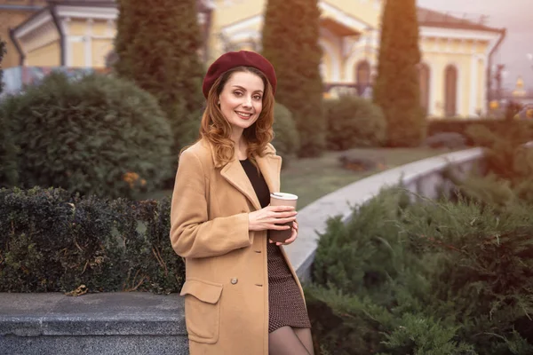 Belle femme parisienne souriante tenant une tasse de café frais debout à l'extérieur beau jardin posant pour la mode féminine. Portrait de la jeune femme élégante portant manteau d'automne et béret rouge — Photo