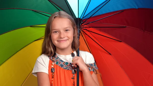 Menina com guarda-chuva de cor arco-íris em suas mãos vestindo vestido laranja de pé feliz sorrindo em estúdio enquanto olha para a câmera — Fotografia de Stock