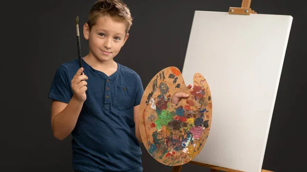 Κρατώντας ένα πινέλο και παλέτα με χρώματα 10χρονο αγόρι σε μπλε t-shirt κοιτάζει την κάμερα στο φόντο ενός καβαλέτου με καμβά. Σχολή Καλών Τεχνών έννοια. Καλλιτεχνική έννοια — Φωτογραφία Αρχείου