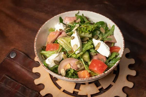 Mísa čerstvého zeleninového salátu s plátky lososa, mozzarella sýr stojí na stole pokrytý koženým hadrem a dekorativní detail jednoduchého mechanismu. Koncept restaurace — Stock fotografie