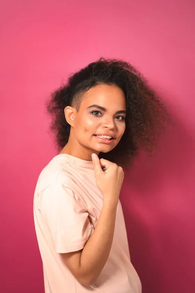 Γοητευτική παθιασμένη νεαρή Αφρο-Αμερικανίδα με τα μαλλιά της να βλέπουν θετικά την κάμερα με το ένα χέρι διπλωμένο φορώντας ένα όμορφο μπλουζάκι απομονωμένο σε ροζ φόντο. Έννοια ομορφιάς — Φωτογραφία Αρχείου