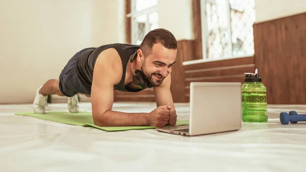Trener fitness robi deskę na podkładce matowej z laptopem podczas wideokonferencji online z klientem podczas samodzielnej izolacji. młody człowiek robi specjalne ćwiczenia dla mięśnie brzucha rdzenia — Zdjęcie stockowe