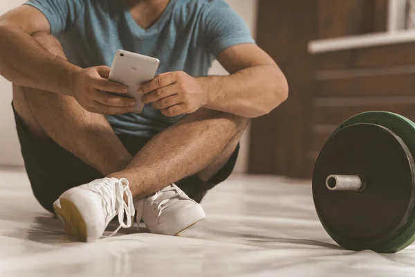 Ninguna foto del cuerpo de un entrenador de fitness buscando en línea o mensajes de texto sentado en un piso con teléfono inteligente, barra de fitness de tono negro y verde, equipo para el concepto de entrenamiento con pesas. Imagen tonificada — Foto de Stock