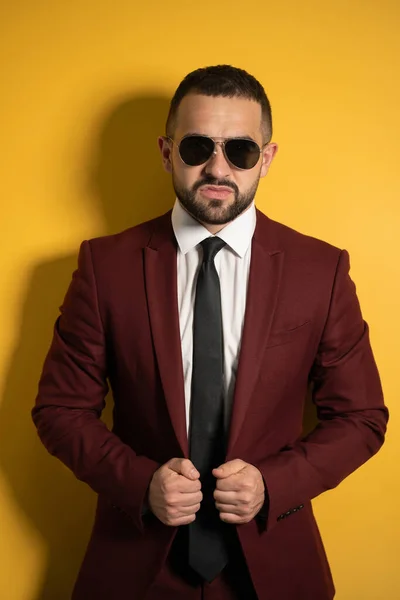 Young handsome man in burgundy suit looking on you seriously wearing sunglasses with hands holding a jacket isolated on yellow background. Men fashion concept — Stock Photo, Image
