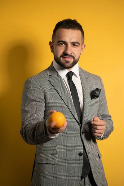 Schöner eleganter Mann im grauen Anzug mit Brille streckt seine Hand aus, in der einen Hand orangefarben, isoliert auf gelbem Hintergrund stehend. Männermode — Stockfoto