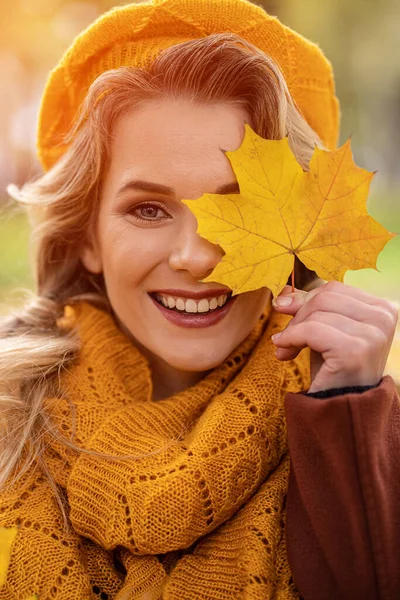 Giovane ragazza felice coprire l'occhio con foglie ingiallite in giallo basco lavorato a maglia con foglie autunnali in mano e autunno giardino giallo o parco. Bella giovane donna sorridente in fogliame autunnale — Foto Stock