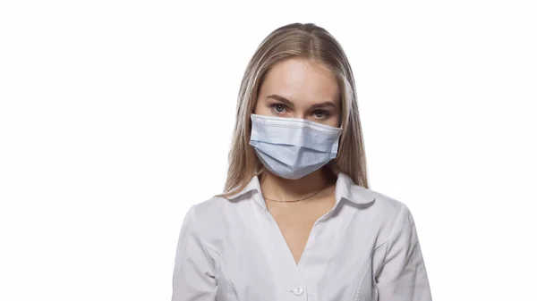 Looks sullenly young nurse in a medical mask and white uniform with blond straight hair looking at the camera. Isolated on white background — Stock Photo, Image