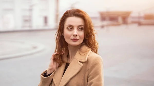 Hermosa mujer de pie al aire libre posando para la cámara en un abrigo beige otoño, hermoso cabello con fondo urbano de la ciudad. Retrato vertical. Foto teñida caliente — Foto de Stock