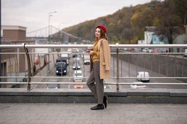 Charmante jeune femme française boit du café à l'aide d'une tasse à café regardant la caméra souriante debout à la passerelle piétonne avec des voitures sur la route. Vous cherchez fille de mode heureuse dans un béret rouge et manteau beige — Photo