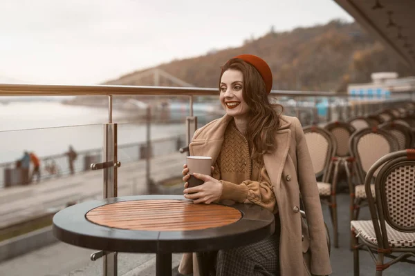 Açık hava lokantasında ya da terasta oturan mutlu genç kız elinde kahve fincanıyla sırıtıyor. Sonbahar ceketi ve açık havada kırmızı bereli şık bir genç kadının portresi. — Stok fotoğraf