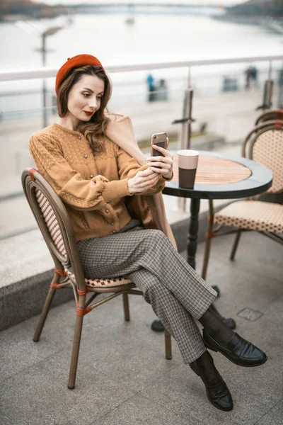 Charmante jeune femme française utilisant son smartphone prenant selfie tout en prenant une tasse de café. Française portant béret rouge assise sur la terrasse d'un restaurant ou d'un café avec fond de ville urbaine d'automne — Photo