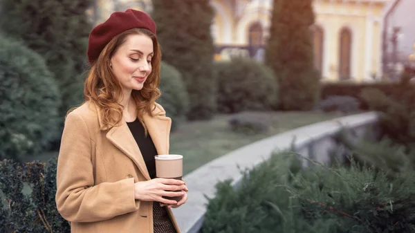 Belle jeune femme tenant une tasse avec du café debout à l'extérieur. Portrait d'une jeune femme élégante portant un manteau d'automne et un béret rouge à l'extérieur. Accessoires d'automne — Photo