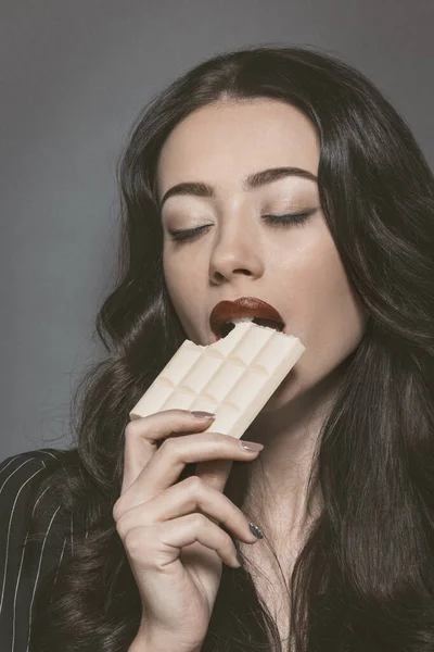 Schöne brünette junge Frau, die weiße Schampus-Schokolade isst. in einer Lederjacke mit schönen langen dunklen Haaren auf grauem Hintergrund. Junges weibliches Model mit professionellem Make-up — Stockfoto