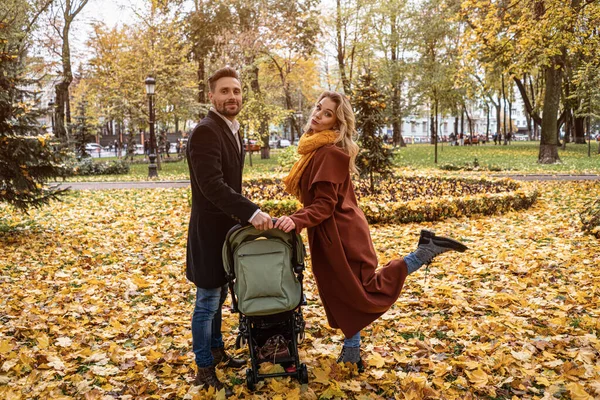 Familia feliz con un bebé recién nacido en un cochecito caminando en un parque de otoño. Feliz madre posando sosteniendo un cochecito con una pierna al aire libre en un parque de otoño dorado. Imagen teñida — Foto de Stock