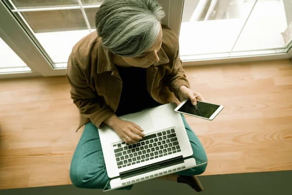 Mulher madura trabalha computador portátil sentado em uma janela. — Fotografia de Stock