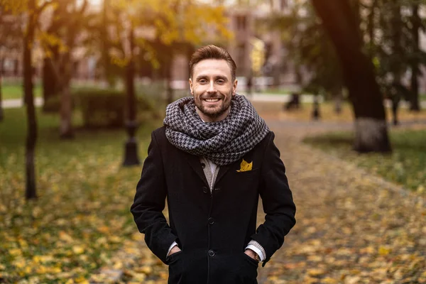 Leende stilig ung man tittar på kameran stående utomhus med händerna i fickorna på sin mörkblå rock. Ung frilansare man står på i en höst rock ser glad ut framför kameran — Stockfoto