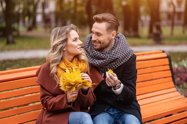 Omfamnande man hittade ett litet löv glada par sitter på bänken kramas i parken bär rockar och halsdukar samla en bukett fallna blad. Kärlekshistoria. Tonad bild — Stockfoto