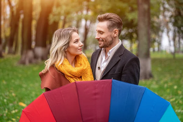 Escondiéndose detrás del paraguas de color arco iris mirándose unos a otros ojos hermosa en pareja de amor de pie en el parque bajo un. Una hermosa pareja el parque de otoño en tiempo lluvioso — Foto de Stock