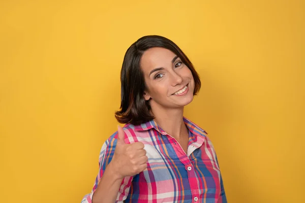 Tillen duim omhoog gebaar met de hand glimlach mooie vrouw gekleed in een geruite shirt en donker haar op gele achtergrond. Menselijke emoties, gezichtsuitdrukking concept — Stockfoto