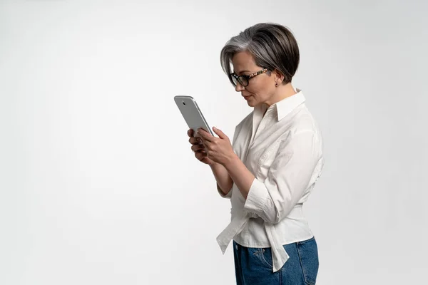 Jolie femme âgée utilisant un ordinateur tablette au bureau en chemise blanche et pantalon en denim. L'utilisation de la technologie par les personnes âgées naviguant sur Internet achats en ligne sur pad isolé sur fond blanc — Photo