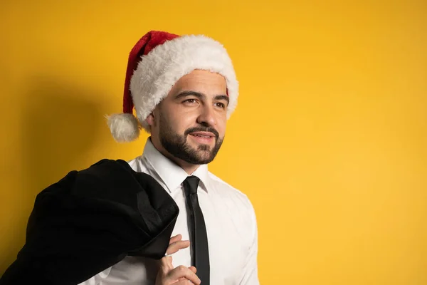 Portret van een knappe Santa zakenman in rode kerstman hoed poseren met zijn zwarte jas op zijn schouder, opknoping het achter kijken vrolijk op camera geïsoleerd op gele achtergrond — Stockfoto