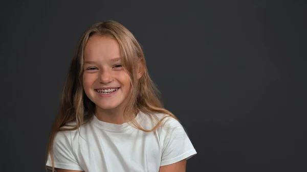 Menina sorridente feliz com cabelo loiro em pé olhando para a câmera vestindo camiseta branca isolada em fundo cinza escuro ou preto — Fotografia de Stock