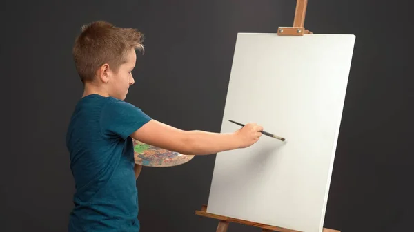 Наносит штрих на холст, держа палитру красок, 10-летний мальчик в синей футболке смотрит в камеру на фоне мольберта с холстом. Концепция школы искусств. Концепция искусства — стоковое фото