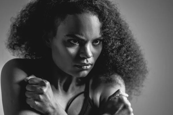 Black and white image. Self-defense, fearful African American girl standing covered with hands in black bare shoulder top isolated on grey background. Human emotions, facial expression concept