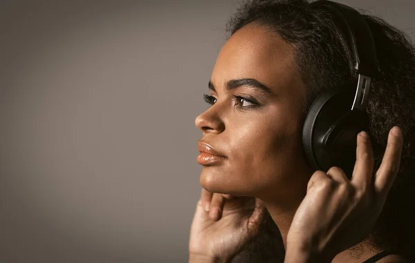 Charmig flicka porträtt av en afroamerikan tittar i sidled lyssna musik bär hörlurar hålla den med händerna, isolerade på grå bakgrund, känslomässigt flytta, ha kul. Närbild — Stockfoto