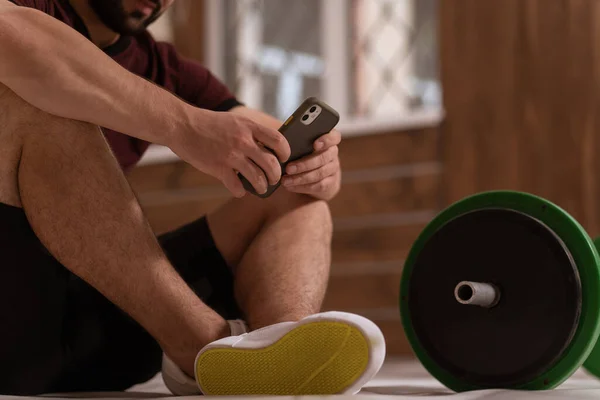 Hombre joven mensajes de texto sentado en un piso con teléfono inteligente, barra de fitness de tono negro y verde, equipo para el concepto de entrenamiento con pesas. Concepto de estilo de vida saludable. Ningún disparo en la cara — Foto de Stock