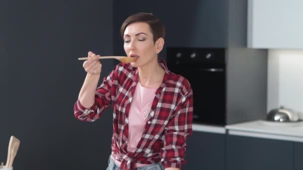 Mulher sexy com um penteado curto testando uma comida enquanto cozinha um jantar para a família com os olhos fechados no prazer em pé na cozinha moderna. Comida saudável em casa. Imagens de FHD — Vídeo de Stock