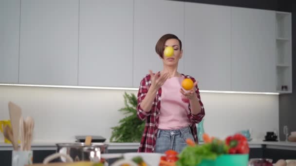 Glückliche Mutter jongliert mit frischer gelber Tomate und Orange in der Hand im karierten Hemd mit Kurzhaarfrisur in der Küche. Gesunde Ernährung zu Hause. Konzept für gesunde Ernährung. — Stockvideo