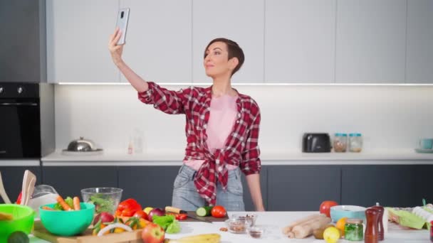 Jolie femme faisant selfie ou appel vidéo à l'aide de son smartphone le tenant dans le bras tendu, tout en cuisinant une salade fraîche portant une chemise à carreaux avec une coiffure bob. Vidéos FHD — Video