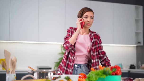 Ter uma conversa no telefone jovem dona de casa bonita com um penteado bob prepara comida na cozinha. Comida saudável em casa. Partida de comida sã - conceito vegan. Imagens de FHD — Vídeo de Stock