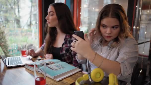 Las niñas estudian la lectura de libros y el uso de un ordenador portátil sentado en un café con luces corriendo y los clientes caminando en el fondo mientras se sientan en una mesa con diferentes libros en ella. Imágenes 4K — Vídeos de Stock