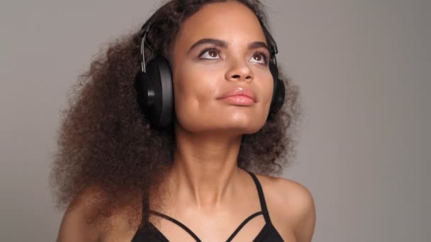 Charmig afroamerikansk tjej tittar upp lyssna musik bär hörlurar blundar i njutning, isolerad på grå bakgrund, känslomässigt flytta, ha kul. 4K-bilder — Stockvideo