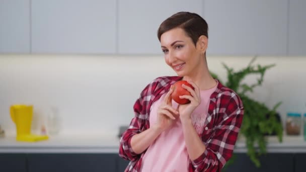 Przetarg gospodyni domowa pachnące świeże jabłko trzymając go w ręce noszenie koszuli w kratę z krótką fryzurę podczas gotowania szarlotki stojącej w kuchni. Zdrowe jedzenie w domu. Nagrania FHD — Wideo stockowe