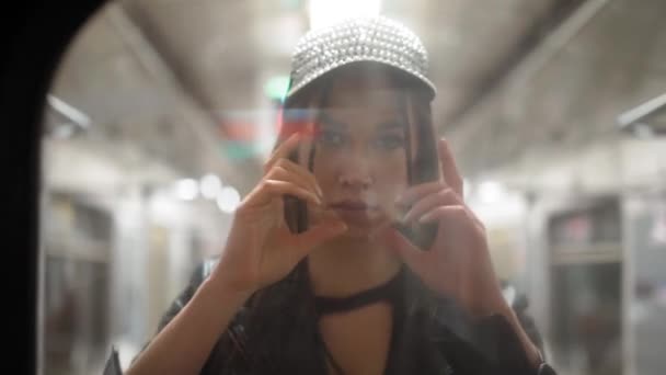 Мода модель позирует через стекло на камере едет в вагоне метро. Красивая девушка выпрямляет волосы и кепку, глядя в зеркало вагона метро. FHD видео — стоковое видео