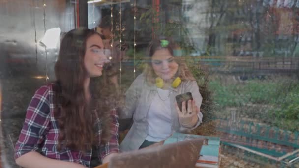 Uitzicht door glas twee vriendinnen selfie terwijl ze samen genieten van koffie in een coffeeshop met klanten lopen op de achtergrond als ze zitten aan een tafel chatten en lachen. 4K-beelden — Stockvideo