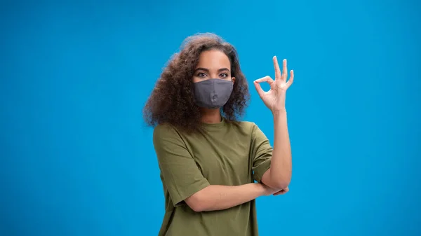 Dizendo OK menina com gesto mulher afro-americana olhando positivamente para a câmera vestindo camiseta verde-oliva com os braços dobrados isolado no fundo azul. Conceito de beleza — Fotografia de Stock