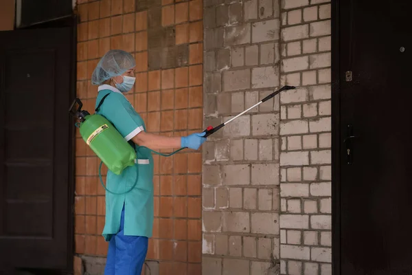 Mujer en traje protector usando contenedor verde para desinfectar la pared exterior del edificio. Tratamiento médico preventivo de la construcción con antiséptico. Hospital Municipal. mayo, 2020, Brovary, Ucrania —  Fotos de Stock