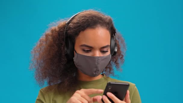 Choisir le prochain titre African American girl écouter de la musique à l'aide d'un smartphone avec casque en t-shirt olive, masque réutilisable, pour prévenir l'infection corona COVID-19 isolé sur fond bleu. 4K. — Video