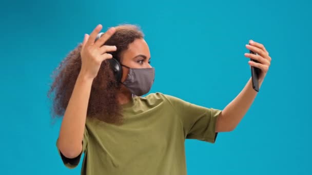 Dancing Afro-Amerikaans meisje luisteren muziek op haar smartphone met koptelefoon in olijf t-shirt, herbruikbaar gezichtsmasker, om te voorkomen dat anderen van corona COVID-19 infectie geïsoleerd op blauwe achtergrond. 4K — Stockvideo