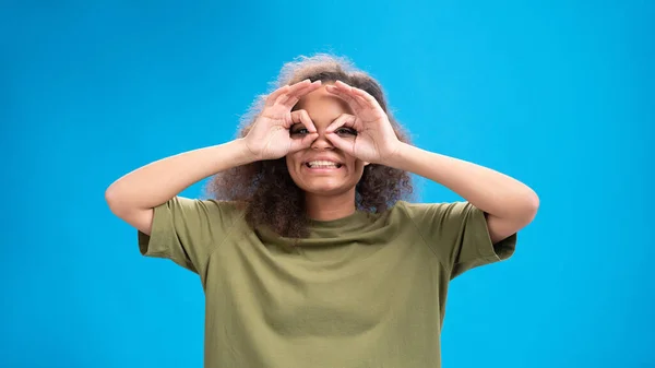 Gesto piloto engraçado menina afro-americana olhando positivamente para a câmera vestindo camiseta verde-oliva com as mãos como binóculos isolados em fundo azul. Conceito de beleza — Fotografia de Stock