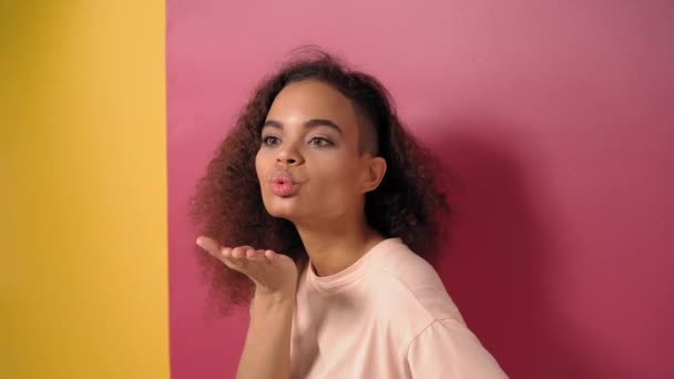 Skickar luft kysser vackra unga afroamerikanska kvinna med afro hår, ser positivt på kameran bär persika t-shirt isolerad på rosa och gul bakgrund. Skönhetskoncept. 4K-bilder — Stockvideo