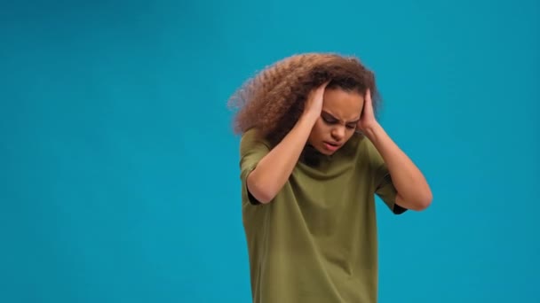 Ощущение мигрени или головной боли афро-американская молодая женщина стоит держа голову с руками в оливковой футболке изолированы на синем фоне. Человеческие эмоции, концепция выражения лица — стоковое видео