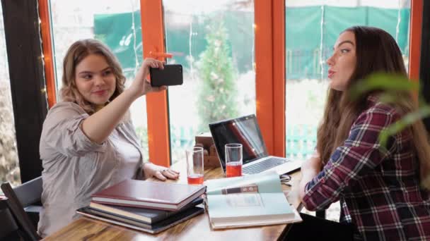 Twee vriendinnen die samen koffie drinken in een koffieshop en boeken lezen om te studeren en laptop met tuin en straat op de achtergrond terwijl ze aan een tafel zitten te kletsen en lachen. 4K-beelden — Stockvideo