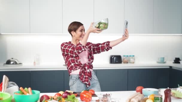Feliz dona de casa fazendo selfie usando seu smartphone segurando uma salada fresca em mão estendida, enquanto cozinha na cozinha moderna em casa vestindo uma camisa xadrez. Imagens de FHD — Vídeo de Stock